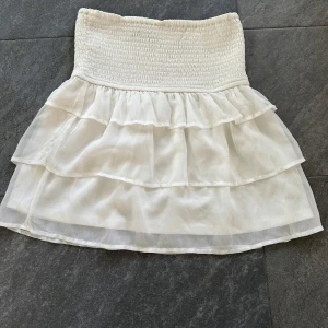 Vit volangkjol med resår - Supersöt vit kjol med bred smock i midjan och dubbla volanglager. Kjolen är kort och har en luftig känsla, perfekt för varma dagar. Passar till både enkla och mer uppklädda outfits.