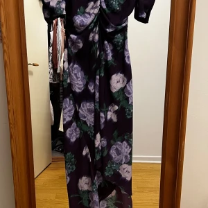 Långklänning med blommigt mönster - Säljer en långklänning med lila och gröna blommor på mörklila botten. Klänningen har puffärmar och draperad detalj framtill. Perfekt för dig som vill sticka ut med ett romantiskt och elegant plagg.