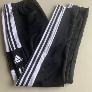 Två svarta Adidas mjukisbyxor med vita ränder - Två svarta mjukisbyxor från Adidas med klassiska vita ränder längs benen och Adidas-logga på ena benet. Resår i midjan och smal passform. Perfekta för träning eller chill.