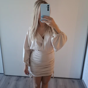 Champagnefärgad klänning från Bubbleroom - Snygg kort klänning från Bubbleroom i champagnefärgad ton. Omlottmodell med v-ringning, ballongärmar och snörning i sidorna som ger en cool rynkad effekt.