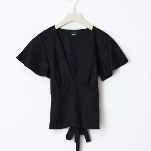 Svart topp från Gina ”tie top”  - Svart topp från Gina tricot med djup v-ringning och korta, fladdriga ärmar. Toppen har knytband i midjan som ger en snygg siluett. Tillverkad i mjuk bomull, perfekt att styla med jeans eller kjol för en chill look. Skicka för mer bilder säljer för endast 180 är helt ny kostar 279 ny😇