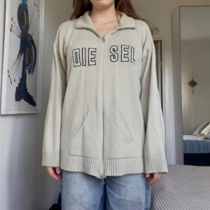 Beige stickad kofta från Diesel - Säljer en beige stickad kofta från Diesel med svart broderad logga på bröstet. Koftan har dragkedja framtill, ribbade muddar och krage. Långärmad och med ficka framtill.