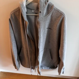 Grå hoodie från Nicki Studios - Säljer en grå hoodie från Nicki Studios med dragkedja, huva och snörning nertill. Tröjan har två stora fickor framtill och är fodrad med mjukt material på insidan. Perfekt för alla dagar och har en clean look med diskret logga på bröstet.                Nypris 1200kr, säljes för 500kr. Kom med prisförslag