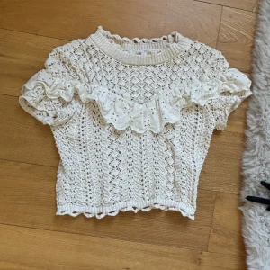 Vit stickad blus med volang Zara - Supersöt vit stickad blus från Zara med korta ärmar och volangdetalj över bröstet och axlarna. Blusen har ett vackert hålmönster och ribbad halsringning. Perfekt för dig som gillar romantisk och vintage-inspirerad stil.