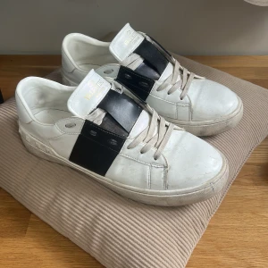 Valentino Garavani Open sneakers vit/svart - Snygga Valentino Garavani Open sneakers i vitt skinn med bred svart läderdetalj över vristen och ikoniska nitar baktill. Klassisk rund tå, platt sula och snörning framtill. Perfekta för dig som gillar lyxiga och stilrena sneakers.