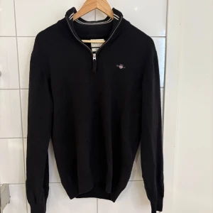 Svart half zip tröja från GANT - Svart långärmad tröja från GANT i superfin lammull. Klassisk half zip med ribbad krage och diskret logga på bröstet. Perfekt för dig som gillar stilrena och tidlösa plagg. Snygga vita ränder på kragen ger extra detalj. Passar dig som vill ha en normal passform.