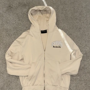 Beige hoodie Supply & Demand XS - Beige hoodie från Supply & Demand med dragkedja och huva. Tryck fram med texten 'supply New York City' och stor grafisk ängel och text på ryggen. Mjuk bomullskänsla och känguruficka framtill. Perfekt för en streetwear-look.