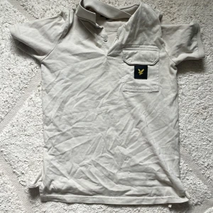 Beige Lyle & Scott t-shirt med ficka - Snygg beige t-shirt från Lyle & Scott med klassisk krage och korta ärmar. På bröstet finns en ficka med det ikoniska Lyle & Scott-märket i svart och gult. Tillverkad i mjuk bomull, perfekt för chill dagar. Passar dig som gillar stilren och enkel design.