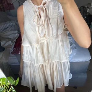 Champagnefärgad omlottklänning med volanger - Säljer en superfin kortklänning i champagnefärgad ton med omlott och volangdetaljer. Klänningen har ett snyggt knyt vid halsen och är ärmlös. Materialet är glansigt och lätt, perfekt för sommarens alla tillfällen. Passformen är loose och ger en skön och luftig känsla.