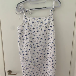 Vit blommig kortklänning från Missguided - Supersöt vit kortklänning från Missguided med blått blommigt mönster. Klänningen har smala axelband med knyt och en rak, loose passform. Perfekt för sommarens alla tillfällen! Materialet är mjukt och lätt polyester.