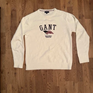 Vit stickad tröja från GANT - Säljer en snygg vit tröja från GANT i storlek L och i bra skick. Den har ett coolt tryck med amerikansk flagga och texten 'New Haven Conn. USA'. Tröjan är långärmad och har ribbade muddar vid ärmslut och nederkant. 