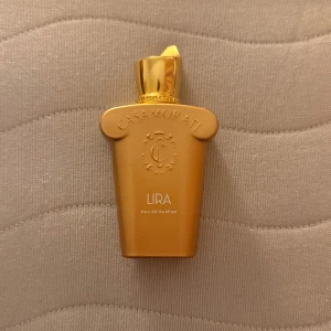 Lira Eau de Parfum från Casamorati - Exklusiv parfymflaska i guldigt material från Casamorati, modell Lira. Flaskan har en unik, lyxig form med präglad logotyp och elegant lock. Perfekt för dig som vill sticka ut med en sofistikerad doft.