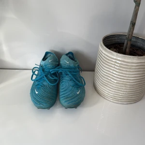 Blå Nike fotbollsskor med snörning - Säljer ett par blå Nike Phantom fotbollsskor med snyggt mönstrad ovandel och vita swoosh-loggor. Skorna har snörning och plös i samma färg, samt dobbar för gräsplan. Perfekta för dig som vill sticka ut på planen. Skorna är använda lite men inget tecken på skada på skorna. Dem fungerar helt. 