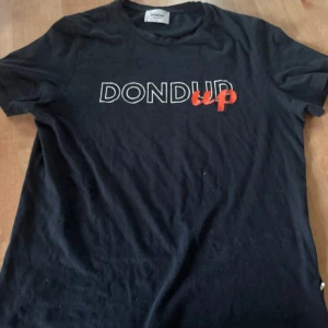 Svart t-shirt från Dondup - Svart t-shirt från Dondup med vit och röd textprint på bröstet. Klassisk rund hals och korta ärmar. Materialet är mjuk bomull som känns skönt mot huden. Snygg och enkel design med coolt tryck, perfekt för dig som gillar italienska märken.