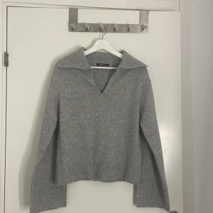 Grå stickad tröja från Gina Tricot - Supermjuk grå stickad tröja från Gina Tricot med v-ringning och bred krage. Tröjan har långa ärmar och ribbade muddar vid ärmslut och nederkant. Perfekt för lager-på-lager och chill höst- och vinterstil.