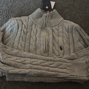 Grå stickad polotröja från Polo Ralph Lauren - Säljer en grå stickad polotröja från Polo Ralph Lauren med klassiskt flätmönster och half zip framtill. Tröjan har hög krage, långärmade ribbade muddar och den ikoniska Polo-loggan broderad på bröstet. Perfekt för dig som gillar stilren och tidlös design.