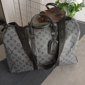 Keepall grå weekend väska - Säljer en grå weekend väska Keepall med klassiskt monogrammönster i svart. Väskan har både handtag och justerbar axelrem med logga, samt detaljer i svart skinn. Perfekt för weekendresor och riktigt snygg att bära med sig.