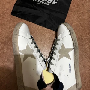Golden Goose vita sneakers med stjärna - Golden Goose sneakers i vitt skinn med grå mockadetaljer och ikonisk stjärna på sidan. Beige snörning, svart hälparti med logga och slitstark platt sula. Snygg kontrast mellan material och färger, perfekt för dig som gillar streetstyle med premiumkänsla.