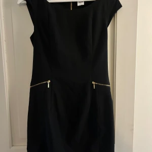 Svart kort klänning från Vero Moda - Svart kort klänning från Vero Moda med snygga guldiga dragkedjor både bak och vid fickorna. Stilren och figurnära modell med korta ärmar och rund halsringning. Perfekt för dig som gillar en enkel men cool look.
