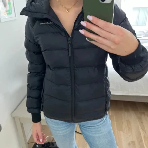 Svart dunjacka Peak Performance - Säljer en svart dunjacka från Peak Performance med huva och dragkedja framtill. Jackan är quiltad och har en clean look med diskret logga på bröstet. Perfekt för kalla dagar och enkel att styla med jeans eller hoodies.
