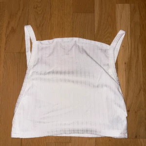 Vit ribbad linnetopp med smala axelband - Fräsch vit linnetopp med smala axelband och diskret ribbad struktur. Toppen är ärmlös och har en enkel, rak form som gör den lätt att matcha med olika outfits. Perfekt för varma dagar och chill stunder med vänner.
