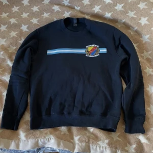 Svart DIF sweatshirt med tryck - Svart sweatshirt med Djurgårdens IF-emblem på bröstet och blåvit rand över bröstet. Klassisk rund hals och långa ärmar. Perfekt för dig som vill visa ditt stöd för DIF på ett stilrent sätt.