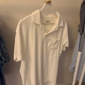 Vit frotté t-shirt från Stones - Vit t-shirt från Stones i mjuk frotté med klassisk krage och korta ärmar. Har en liten bröstficka som ger en chill vibe. Perfekt för dig som gillar clean och enkel stil med lite retrokänsla. Passformen är normal och materialet känns skönt mot huden.