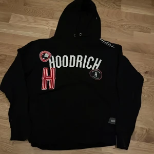 Svart hoodie från Hoodrich med tryck - Svart hoodie från Hoodrich med stor vit logotyp och röda detaljer på bröstet. Tröjan har huva med snörning och långa ärmar. Materialet är mjuk bomull och passformen är normal. Perfekt för dig som gillar streetwear och vill sticka ut med coola tryck.