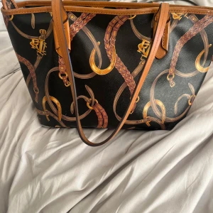 Svart axelväska från Lauren Ralph Lauren - Snygg svart axelväska från Lauren Ralph Lauren med brunt och guldigt mönster av hästbetsel och flätade detaljer. Väskan har bruna handtag, dragkedja med RLL-logga och randigt innerfoder i beige och svart. Perfekt storlek för allt du behöver ha med dig.