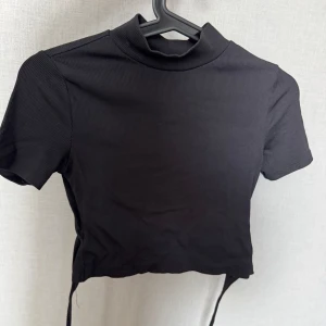 Svart croppad polotopp med snörning i bak  - Svart ribbad polotopp med korta ärmar och croppad passform. Toppen har  snörning i bak och en öppen rygg. 