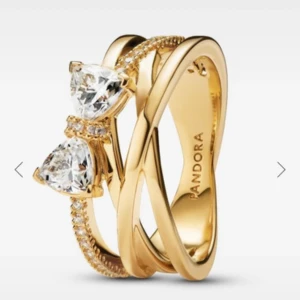 Guldfärgad ring med stenar från Pandora - Superfin guldfärgad ring från Pandora med två stora, gnistrande hjärtformade stenar och små glittriga stenar längs ena bandet. Ringen har en cool, korsad design som verkligen sticker ut och ger en lyxig känsla.