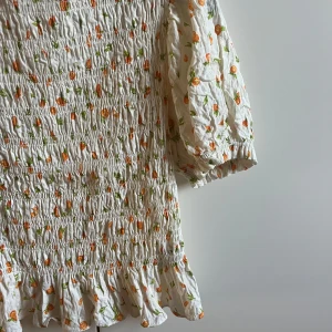 Vit blommig fintröja från H&M - Supersöt fintröja från H&M Divided i vitt med orange och grönt blommigt mönster. Den har puffiga trekvartsärmar, smockad överdel och volangkant nedtill. 