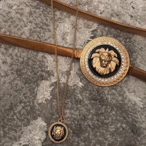 Halsband och bälte - Säljer ett smalt guldigt bälte i stretchigt one size med ett stort runt spänne prytt med ett lejonhuvud och gnistrande stenar runtom. Spännet har svart bakgrund och grekisk-inspirerat mönster. Bältet är i metall och har en riktigt lyxig vibe. Matchande halsband till. Bältet är köpt på FashionNova och halsbandet är från Glitter.