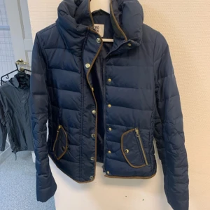 Marinblå pufferjacka med detaljer - Snygg marinblå pufferjacka från Zero Moda med bruna kantdetaljer och guldiga knappar. Jackan har dragkedja, tryckknappar och två fickor framtill. Perfekt för kyliga dagar och har en hög krage som skyddar mot vinden.