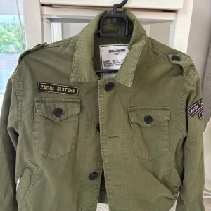 Grön overshirt från Zadig & Voltaire - Snygg militärgrön overshirt från Zadig & Voltaire med coola badges som 'Zadig Sisters' och 'Keep the dream alive'. Jackan har knappar framtill, axelklaffar och två bröstfickor. Perfekt för dig som vill ha en streetig och avslappnad stil. Nästan aldrig använt