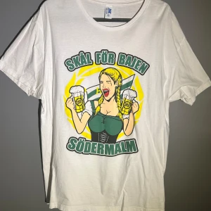 Vit Bajen t-shirt med tryck - Vit t-shirt med stort färgglatt tryck på framsidan: en tecknad tjej med flätor, två ölglas och texten 'Skål för Bajen Södermalm' i grönt och gult. Rund hals och korta ärmar. Perfekt för dig som älskar Bajen och Södermalm.