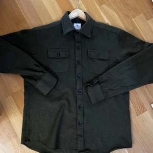 Olivgrön overshirt från John Henric - Snygg olivgrön overshirt från John Henric med klassisk krage, två bröstfickor med lock och knappar framtill. Skjortan är i ett mjukt material som känns lite tjockare, perfekt att ha som lager. Långärmad och stilren design som funkar till många outfits. Storlek L men sitter som en M. Nypris 999kr.