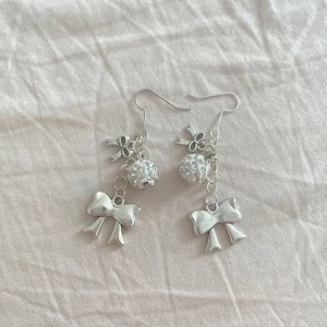 Silvriga örhängen med rosetter och pärlor - Supersöta silvriga örhängen med hängande rosetter och glittrande pärlor. De har små detaljer i form av rosetter och en rund pärla med strass som ger extra bling. Perfekta för dig som gillar gulliga accessoarer med lite extra sparkle.