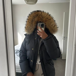 Svart dunjacka med äkta päls från Boomerang - Säljer en svart dunjacka från Boomerang med stor fluffig huva kantad av äktapäls. Jackan har dragkedja, knappar och dragsko i midjan. Yttermaterial i polyester och fyllning av dun och fjäder. Perfekt för kalla dagar och riktigt snygg passform.