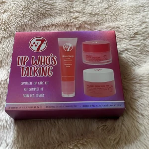 W7 Lip Who's Talking läppkit - Säljer ett komplett läppvårdskit från W7 med tre produkter: ett glossigt läppbalsam, en söt läppskrubb och en overnight läppmask, alla med jordgubbssmak. Förpackningen är rosa och lila med tydliga produktbilder. Perfekt för dig som vill ha mjuka och glansiga läppar.