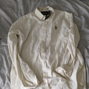 Beige linneskjorta från Lyle & Scott - Snygg beige skjorta från Lyle & Scott i linne med klassisk krage och knappar framtill. Skjortan har lång ärm och den ikoniska gula örnloggan broderad på bröstet. Perfekt för dig som gillar stilrena och avslappnade plagg.