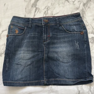 Jeanskjol från U.S. Polo Assn. - Snygg jeanskjol från U.S. Polo Assn. i klassisk blå denim med slitningar och broderad logga vid fickan. Kjolen har fem fickor, bälteshällor och kontrastsömmar i orange. Perfekt till sneakers eller boots för en chill look.