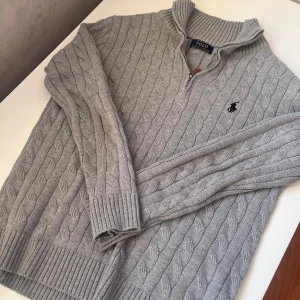 Grå stickad tröja Polo Ralph Lauren - Snygg grå kabelstickad tröja från Polo Ralph Lauren med half zip och ribbad krage. Tröjan har lång ärm och klassisk broderad logga på bröstet. Perfekt för dig som gillar stilren och tidlös design med premiumkänsla.