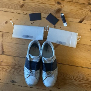 Valentino Garavani sneakers  - Säljer ett par snygga Valentino Garavani sneakers i vitt skinn med bred blå detalj över mitten. Klassisk rund tå, vita snören och platt sula. Skorna kommer med originalkartong, dustbags och extra skosnören. Tveka inte att höra av er om ni har några frågor😊
