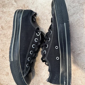 Svarta Converse sneakers med rutigt foder - Svarta Converse sneakers i canvas med vita kontrastsömmar och klassisk snörning. Insidan har ett schackrutigt svartvitt foder som ger en cool detalj. Skorna har rund tå och platt sula, perfekta för en avslappnad streetstil.