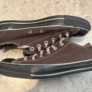 Bruna Converse All Star sneakers - Snygga bruna Converse All Star sneakers i canvas med klassisk låg modell. Skorna har beige snörning, svart tåhätta och svart sula med ljusgrön rand. Insidan har rutigt mönster och tydlig All Star-logga på hälen. Perfekta för en avslappnad streetstil.