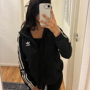 Svart Adidas vindjacka med huva - Svart vindjacka från Adidas med klassiska vita ränder längs ärmarna och broderad logga på bröstet och ärmen. Jackan har huva, dragkedja framtill och är tillverkad i lätt polyester. Aldrig använd. 