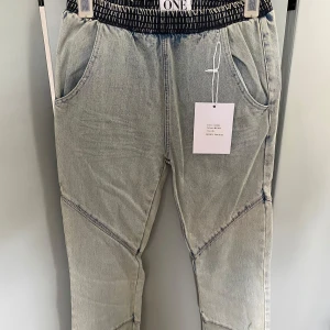 Ljusgrå baggy jeans från ONE teaspoon  - Baggy jeansbyxor från ONE teaspoon i ljusgrå denim med coola sömdetaljer över benen och elastisk midja med svartvitt mönster. Byxorna har fickor fram och bak, samt en avslappnad passform som ger en chill vibe. Storlek 26.