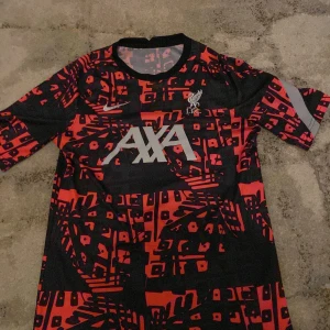 Liverpool tränings-tshirt Nike röd/svart - Liverpool FC tränings-tshirt från Nike med grafiskt rött och svart mönster. Stort AXA-tryck på bröstet, klubbmärke och Nike-logga, perfekt för dig som är fotbollsfan tröjan köptes för 699kr och säljs pga ingen användning 