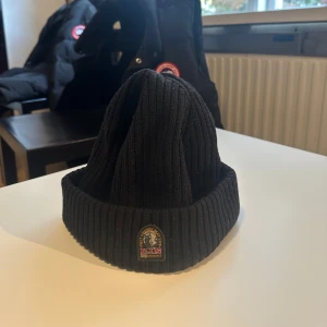 Svart ribbad mössa från Parajumpers - Svart ribbstickad mössa från Parajumpers med uppvikt kant och snyggt broderat märke framtill. Mössan är inte använd så mycket och säljs nu pga den ej passar min stil längre. 